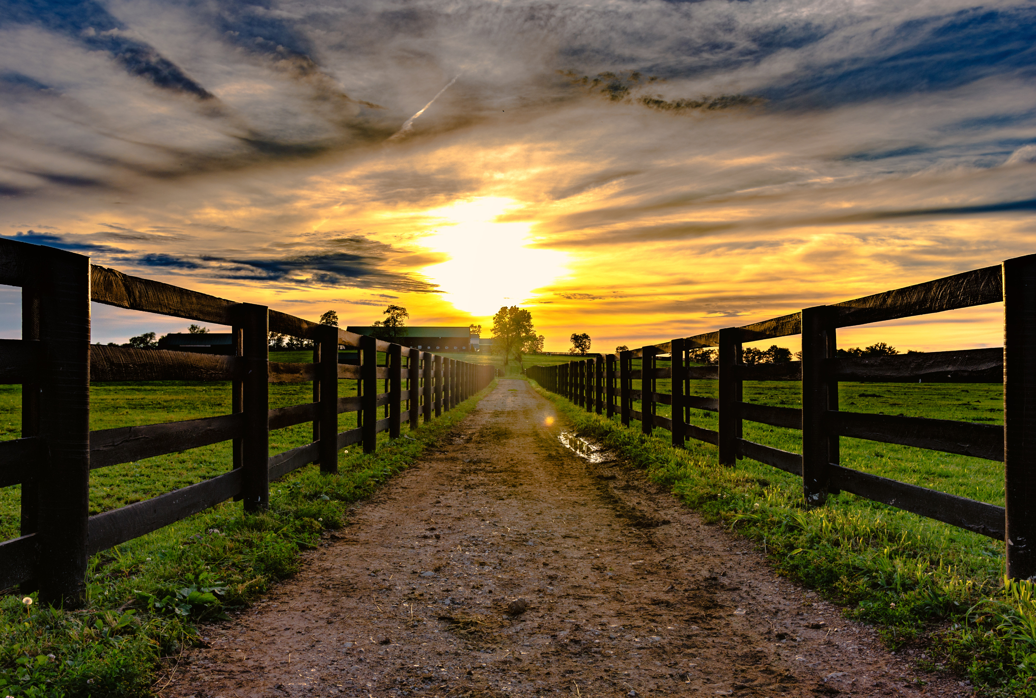 Dirt road leading to a barn in the distance with wooden rail fences on either side with a sunset in the background